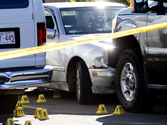 A photo of a white sedan with multiple bullet holes in the windshield and the front bumper compressed into the front of a black van. Numerous yellow evidence markers and police tape surround the scene.
