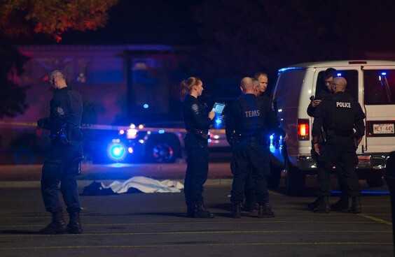 A photo of the crime scene at night, with a white tarp laid over a body on the ground. In the foreground, a group of first responders including police, and TEMS (Tactical Emergency Medical Support) personnel are gathered, next to several police vans.