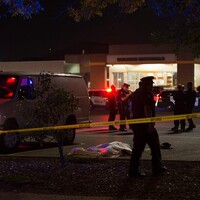 A photo of the crime scene at night, roped off with yellow crime scene tape. A body on the ground is covered by a white tarp; a group of first responders are gathered in the background and a police officer walks past in the foreground.